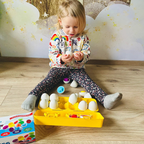 A child playing with a set of Montessori matching eggs on the floor. The set includes multiple colored eggs, and the child is matching them with a set of shapes in a container.
